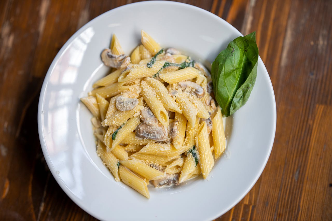 Delicious creamy penne pasta garnished with basil, mushrooms, and parmesan cheese on a rustic wooden table.